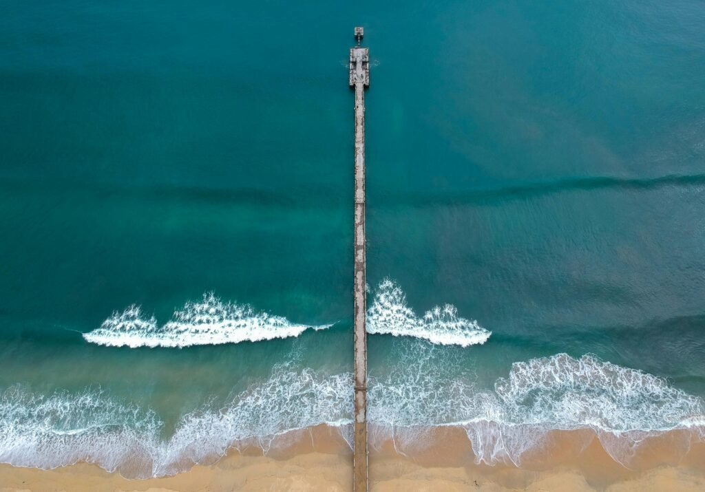 Aerial shot of a long pier extending into the ocean with waves crashing on the sandy shore.